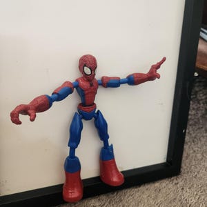 May include: A red and blue plastic action figure of a superhero character with a spider-web pattern on the head and chest. The figure has articulated joints at the shoulders, elbows, hips, and knees, and is posed with arms extended outward.