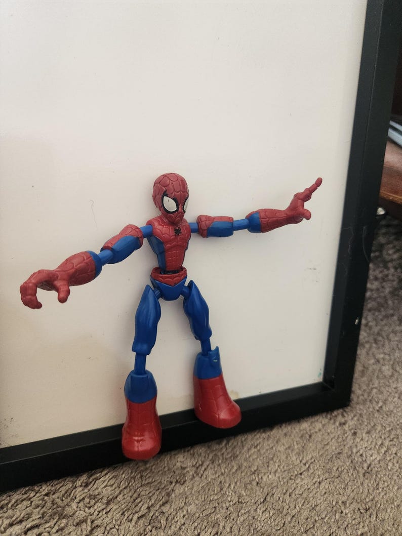 May include: A red and blue plastic action figure of a superhero character with a spider-web pattern on the head and chest. The figure has articulated joints at the shoulders, elbows, hips, and knees, and is posed with arms extended outward.