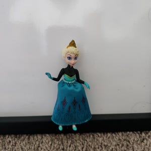 May include: A small plastic doll of Elsa from the movie Frozen. She is wearing a blue and black dress with a gold crown.