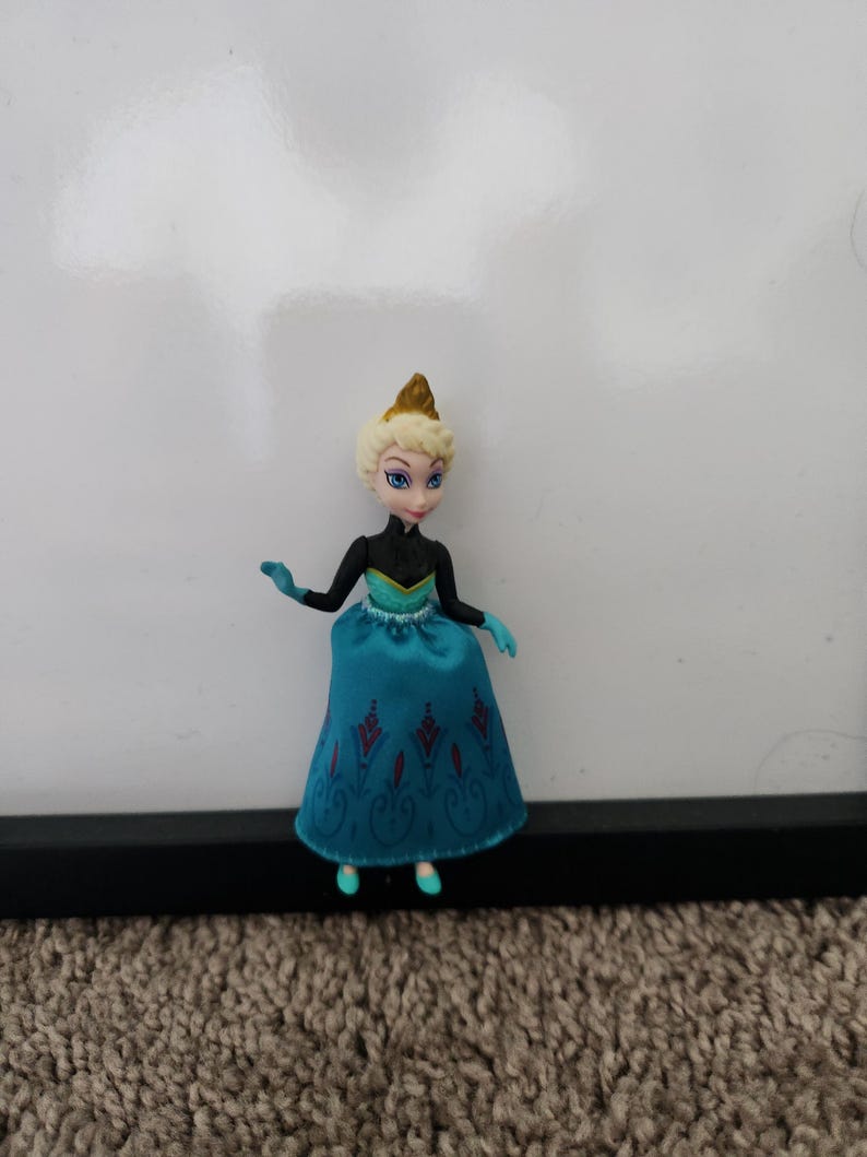 May include: A small plastic doll of Elsa from the movie Frozen. She is wearing a blue and black dress with a gold crown.