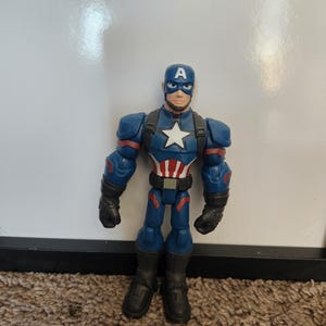 May include: A Captain America action figure. The figure is primarily blue with red and white accents. It features a helmet with a white "A", a white star on the chest, and black gloves and boots. The figure is standing on a brown carpet.