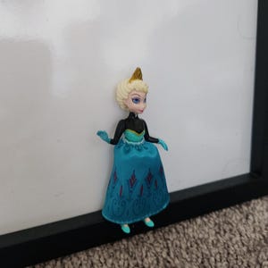 May include: A small plastic doll of a princess wearing a blue and black dress with a gold crown. The doll has blonde hair and blue eyes.