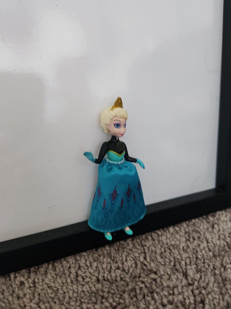 May include: A small plastic doll of a princess wearing a blue and black dress with a gold crown. The doll has blonde hair and blue eyes.
