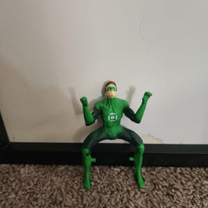 May include: A Green Lantern action figure in a dynamic pose, with arms raised. The figure is predominantly green with black accents, featuring the Green Lantern emblem on the chest. The figure is on a brown carpet with a white background.