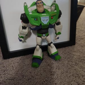 May include: A Buzz Lightyear action figure, standing in front of a white surface. The figure is predominantly green, white, and purple, with a detailed design. The figure has a determined expression and is posed upright.