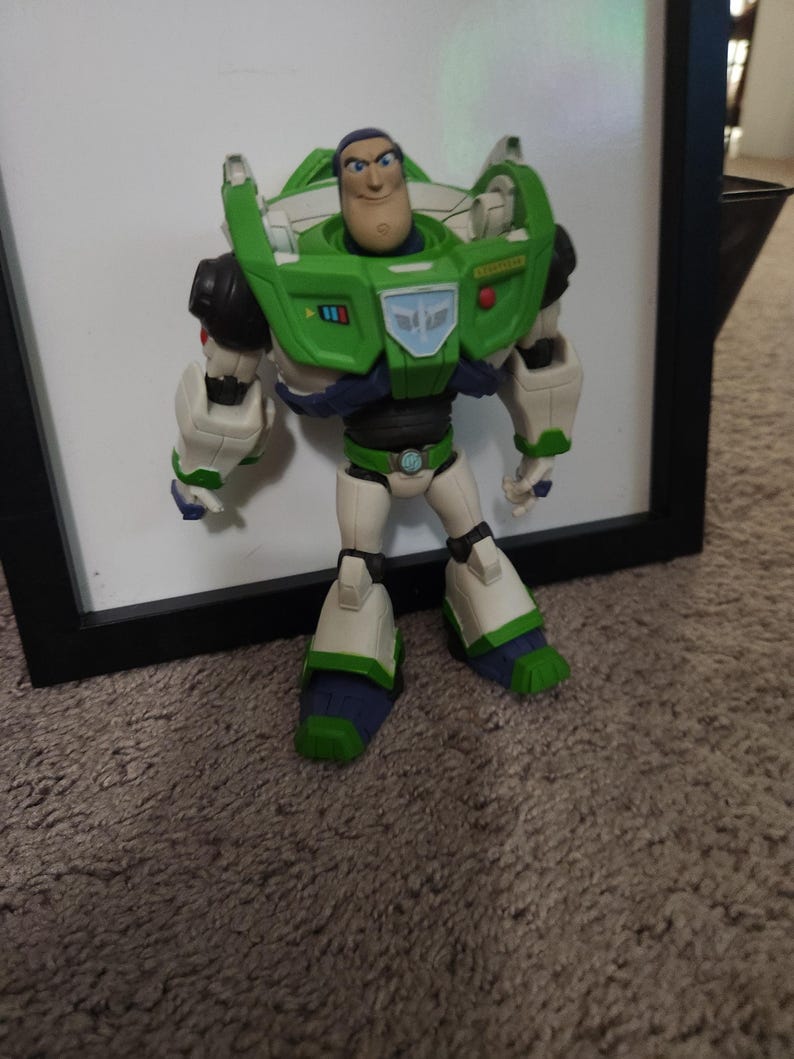 May include: A Buzz Lightyear action figure, standing in front of a white surface. The figure is predominantly green, white, and purple, with a detailed design. The figure has a determined expression and is posed upright.