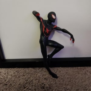 Puede incluir: Figura de acci&oacute;n negra de un superh&eacute;roe en una pose din&aacute;mica. La figura tiene detalles rojos en el pecho, los brazos y las manos. La m&aacute;scara del superh&eacute;roe tiene lentes blancos. La figura est&aacute; sobre un fondo blanco.