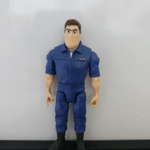 K&ouml;nnte beinhalten: Eine blaue und braune Actionfigur eines Mannes, der einen blauen Overall mit einem wei&szlig;en Hemd darunter tr&auml;gt. Die Figur hat braunes Haar und einen ernsten Gesichtsausdruck.