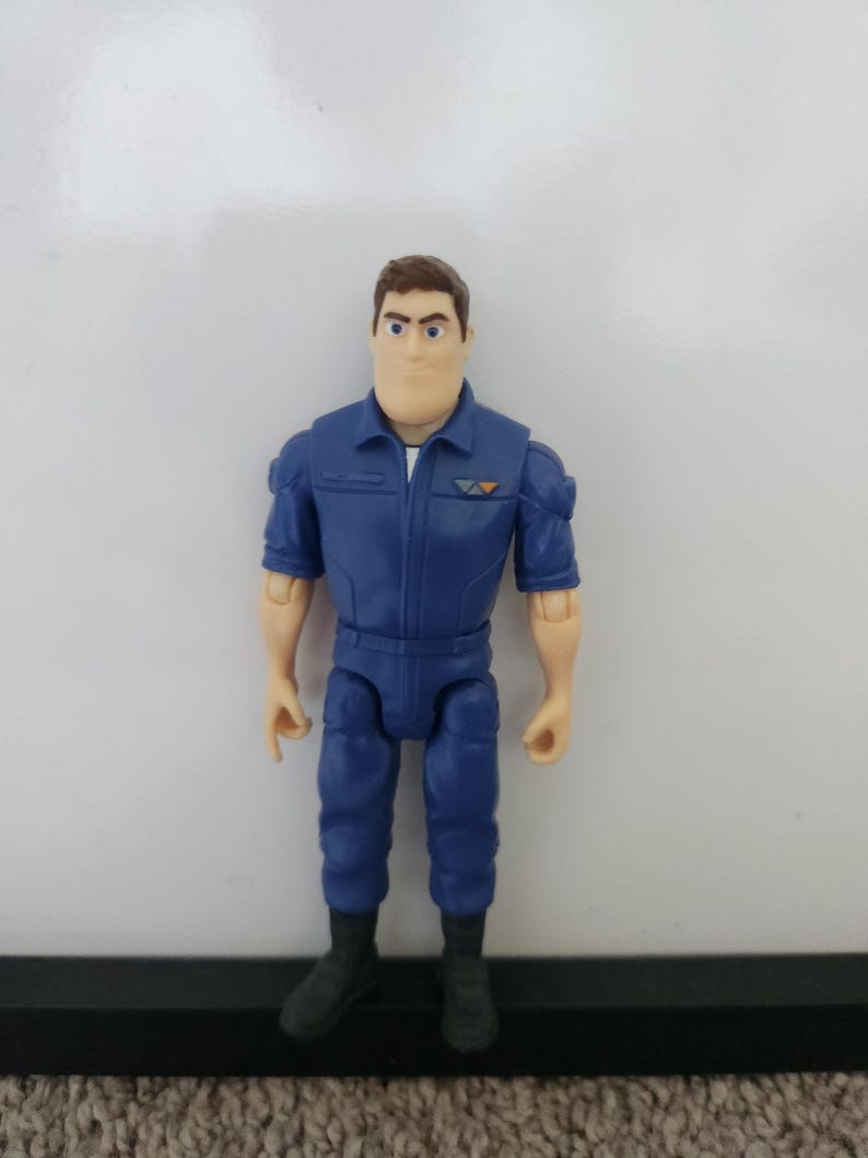 K&ouml;nnte beinhalten: Eine blaue und braune Actionfigur eines Mannes, der einen blauen Overall mit einem wei&szlig;en Hemd darunter tr&auml;gt. Die Figur hat braunes Haar und einen ernsten Gesichtsausdruck.