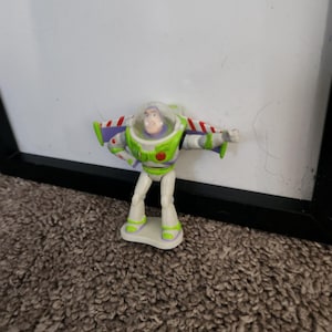 Puede incluir: Una peque&ntilde;a figura de acci&oacute;n de Buzz Lightyear. La figura es principalmente blanca, verde y morada, con un casco transparente y alas rojas y blancas. La figura est&aacute; sobre una peque&ntilde;a base blanca.