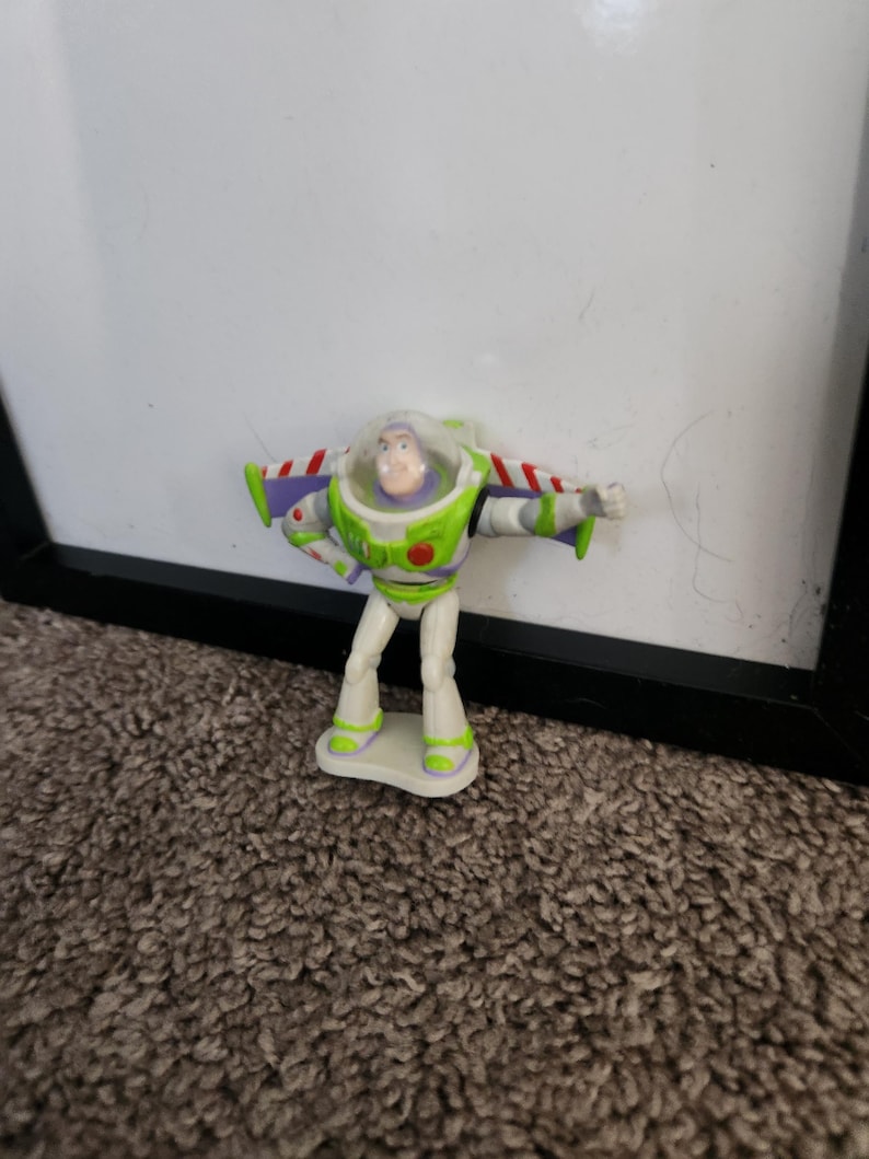 Puede incluir: Una peque&ntilde;a figura de acci&oacute;n de Buzz Lightyear. La figura es principalmente blanca, verde y morada, con un casco transparente y alas rojas y blancas. La figura est&aacute; sobre una peque&ntilde;a base blanca.