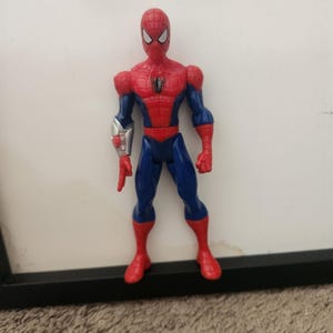 Może przedstawiać: Figurka akcji Spider-Mana w czerwono-niebieskim kostiumie z czarnym emblemem pająka na klatce piersiowej. Figurka posiada srebrny mechaniczny element na prawym przedramieniu i stoi w wyprostowanej pozycji z białymi soczewkami oczu.