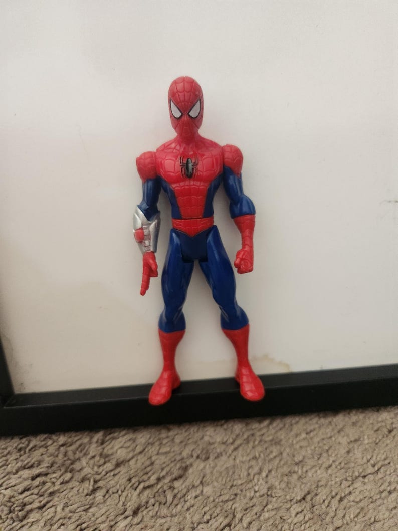 Może przedstawiać: Figurka akcji Spider-Mana w czerwono-niebieskim kostiumie z czarnym emblemem pająka na klatce piersiowej. Figurka posiada srebrny mechaniczny element na prawym przedramieniu i stoi w wyprostowanej pozycji z białymi soczewkami oczu.