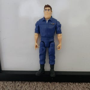 K&ouml;nnte beinhalten: Eine blaue und schwarze Actionfigur eines Mannes, der einen blauen Overall mit einem Aufn&auml;her auf der Brust tr&auml;gt. Die Figur hat braunes Haar und einen ernsten Gesichtsausdruck.