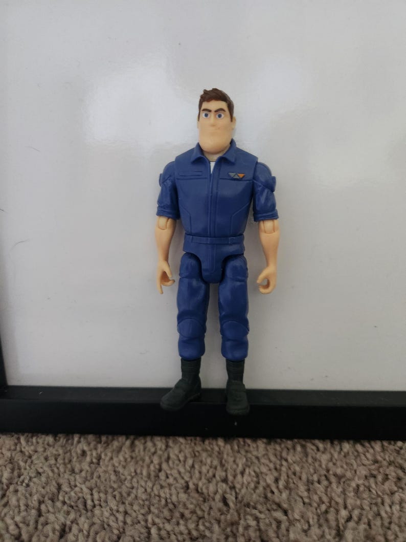 K&ouml;nnte beinhalten: Eine blaue und schwarze Actionfigur eines Mannes, der einen blauen Overall mit einem Aufn&auml;her auf der Brust tr&auml;gt. Die Figur hat braunes Haar und einen ernsten Gesichtsausdruck.