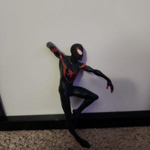 Puede incluir: Figura de acci&oacute;n de Spider-Man negro y rojo en una pose din&aacute;mica. La figura tiene un emblema de ara&ntilde;a roja en el pecho y detalles rojos en los brazos. La figura est&aacute; sobre un fondo blanco.