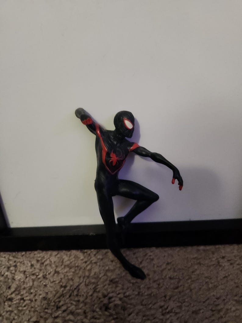 Puede incluir: Figura de acci&oacute;n de Spider-Man negro y rojo en una pose din&aacute;mica. La figura tiene un emblema de ara&ntilde;a roja en el pecho y detalles rojos en los brazos. La figura est&aacute; sobre un fondo blanco.