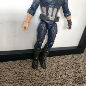 May include: A Captain America action figure. The figure is wearing a blue and gray suit with red and white accents. The figure has black boots and gloves. The figure is standing in front of a white background.
