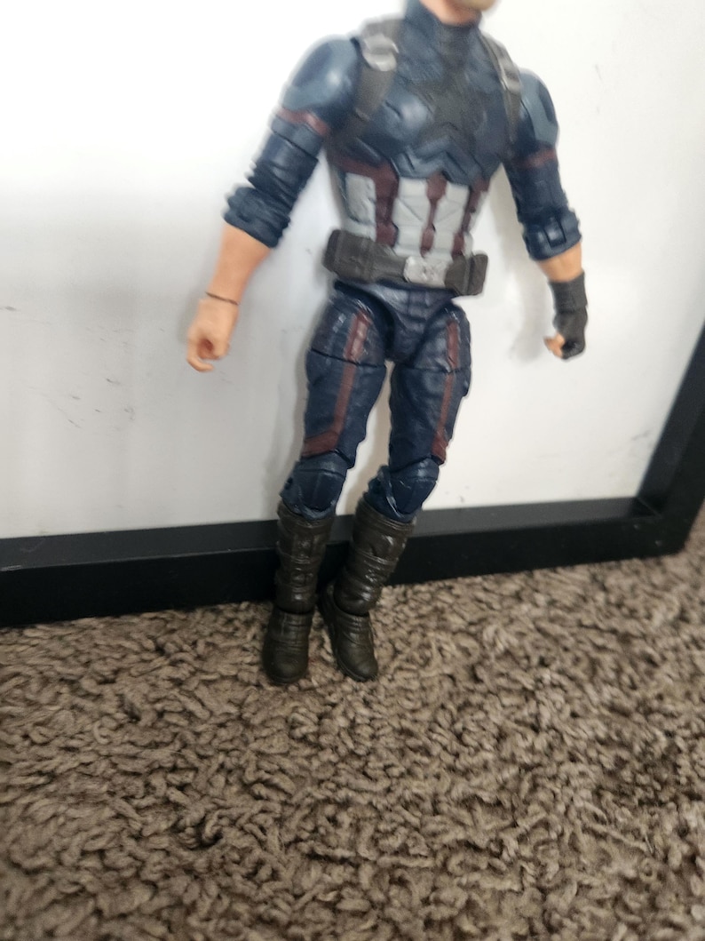 May include: A Captain America action figure. The figure is wearing a blue and gray suit with red and white accents. The figure has black boots and gloves. The figure is standing in front of a white background.