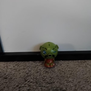 Op de afbeelding: Een groene figuur met een ronde kop, blauwe ogen en een rood-oranje torso. De figuur heeft een klein, rond lichaam en staat rechtop. De achtergrond is een witte muur en een bruin tapijt.