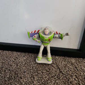 Puede incluir: Figura de acci&oacute;n de Buzz Lightyear de la franquicia Toy Story. La figura es principalmente blanca, verde y morada, con un casco transparente y los brazos extendidos. La figura est&aacute; de pie sobre una peque&ntilde;a base rectangular.