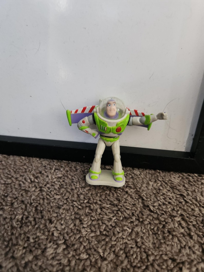 Puede incluir: Figura de acci&oacute;n de Buzz Lightyear de la franquicia Toy Story. La figura es principalmente blanca, verde y morada, con un casco transparente y los brazos extendidos. La figura est&aacute; de pie sobre una peque&ntilde;a base rectangular.