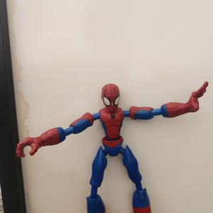 May include: A red and blue plastic action figure of a superhero character with a web pattern on the head and chest. The figure has articulated joints at the shoulders, elbows, hips, and knees, and is posed with arms extended outward to the sides.