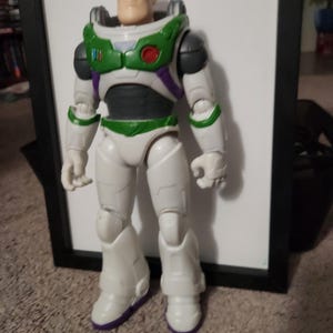 May include: A Buzz Lightyear action figure, featuring a white and green spacesuit with purple accents. The figure has a detailed face and is standing in a neutral pose. The toy is approximately 12 inches tall.