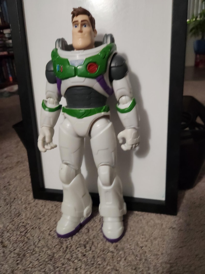 May include: A Buzz Lightyear action figure, featuring a white and green spacesuit with purple accents. The figure has a detailed face and is standing in a neutral pose. The toy is approximately 12 inches tall.