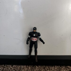 Pu&ograve; includere: Una piccola action figure di Captain America in un abito nero con una stella bianca sul petto. La figura ha un casco nero con la lettera "A" bianca e dettagli bianchi sulle braccia. La figura &egrave; in piedi con una gamba leggermente in avanti.