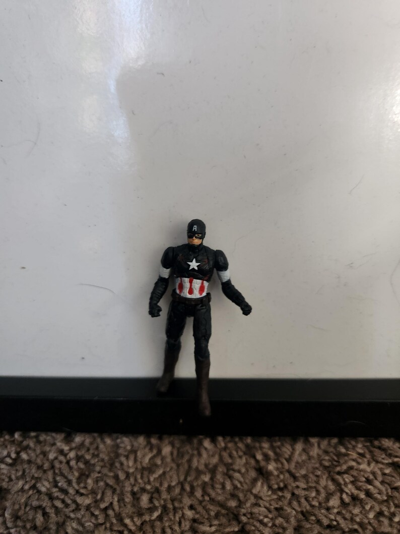 Pu&ograve; includere: Una piccola action figure di Captain America in un abito nero con una stella bianca sul petto. La figura ha un casco nero con la lettera "A" bianca e dettagli bianchi sulle braccia. La figura &egrave; in piedi con una gamba leggermente in avanti.