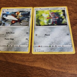 Könnte beinhalten: Zwei Pokémon-Sammelkarten. Die Bisharp-Karte zeigt einen rot-goldenen Charakter. Die Pawniard-Karte zeigt einen kleinen, dunkel gefärbten Charakter mit roten Akzenten. Beide Karten haben gelbe Ränder und Text.
