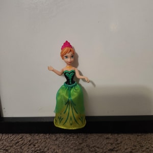 May include: A small doll of a character with orange hair styled in a bun, wearing a green and black dress with gold accents. The doll has a pink crown and is posed with arms outstretched.