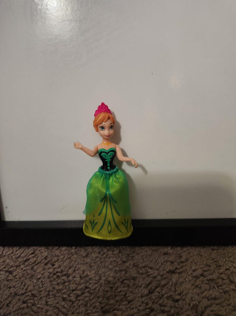 May include: A small doll of a character with orange hair styled in a bun, wearing a green and black dress with gold accents. The doll has a pink crown and is posed with arms outstretched.