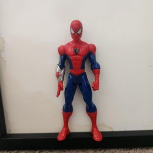 Może przedstawiać: Figurka akcji Spider-Mana w czerwono-niebieskim kostiumie z czarnym emblematem pająka na klatce piersiowej. Figurka ma białe oczy i srebrny dodatek w formie rękawicy na prawym przedramieniu, a prawa dłoń jest ułożona w geście strzelania siecią.