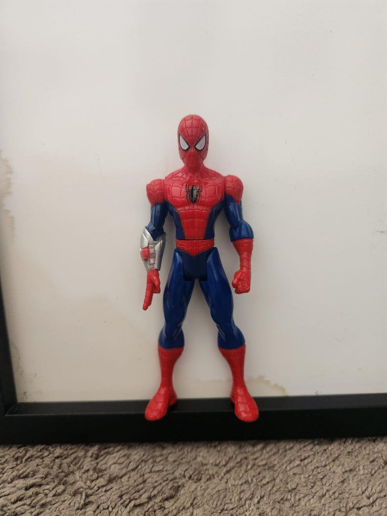 Może przedstawiać: Figurka akcji Spider-Mana w czerwono-niebieskim kostiumie z czarnym emblematem pająka na klatce piersiowej. Figurka ma białe oczy i srebrny dodatek w formie rękawicy na prawym przedramieniu, a prawa dłoń jest ułożona w geście strzelania siecią.