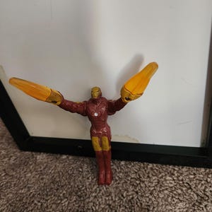 May include: A small Iron Man action figure in a classic pose, with arms outstretched and repulsor rays extended. The figure is primarily red and gold, with yellow energy blasts. The figure is standing in front of a white background.