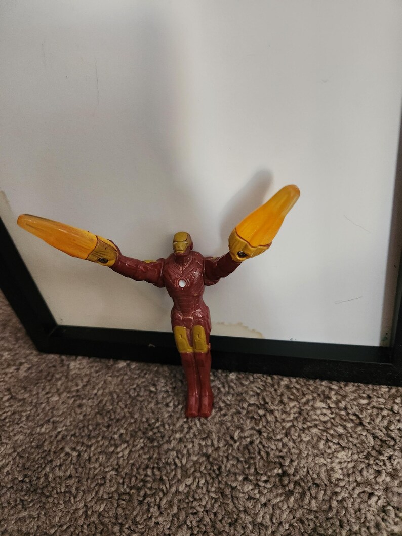 May include: A small Iron Man action figure in a classic pose, with arms outstretched and repulsor rays extended. The figure is primarily red and gold, with yellow energy blasts. The figure is standing in front of a white background.