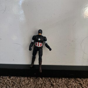 Pu&ograve; includere: Una piccola action figure di Captain America. La figura &egrave; per lo pi&ugrave; nera con accenti bianchi e rossi. Ha una stella bianca sul petto e un casco nero con una "A" bianca. La figura &egrave; su un tappeto marrone.