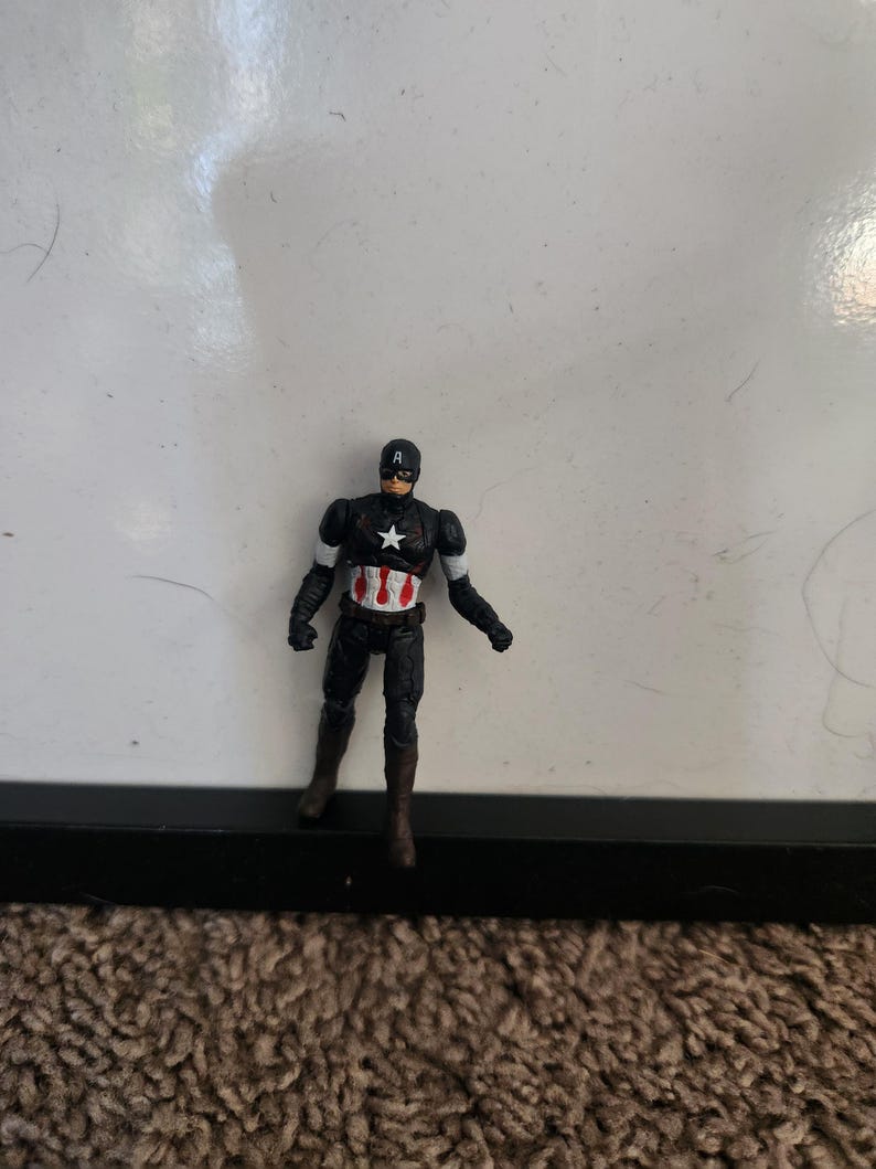 Pu&ograve; includere: Una piccola action figure di Captain America. La figura &egrave; per lo pi&ugrave; nera con accenti bianchi e rossi. Ha una stella bianca sul petto e un casco nero con una "A" bianca. La figura &egrave; su un tappeto marrone.