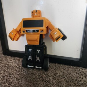May include: A toy robot in orange and black. The robot has a rectangular body with the letters "LYB" printed on the front. The arms and head are orange, and the lower body is black. The toy is standing on a brown surface.
