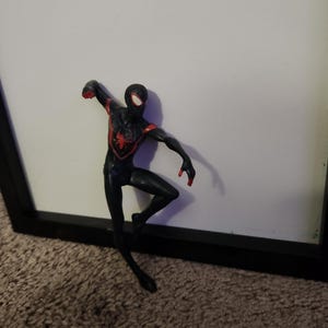 Puede incluir: Figura de acci&oacute;n de Spider-Man negro y rojo en una pose din&aacute;mica. La figura tiene un emblema de ara&ntilde;a roja en el pecho y detalles rojos en brazos y manos. Fondo blanco.