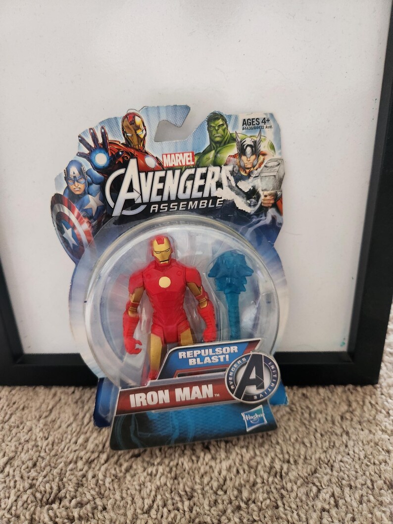 May include: A plastic action figure of Iron Man from the Marvel Avengers Assemble series. The figure is red and gold with a repulsor blast effect. The figure is packaged in a plastic blister pack with a blue and white background. The packaging features the Marvel Avengers Assemble logo and the words "Repulsor Blast!" and "Iron Man".