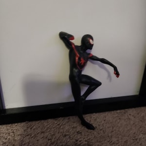 Puede incluir: Figura de acci&oacute;n de Spider-Man negro y rojo en una pose din&aacute;mica, apoyada contra una superficie blanca. La figura tiene un dise&ntilde;o detallado con detalles rojos en el pecho, los brazos y la m&aacute;scara. Aproximadamente 15 cm de altura.