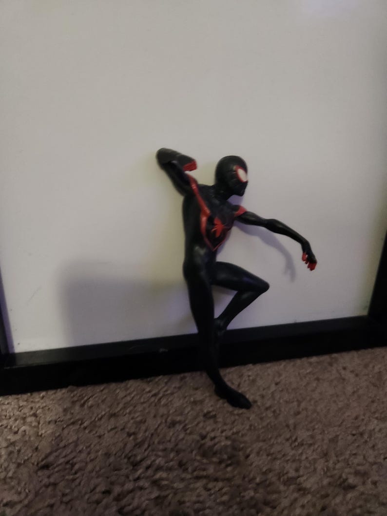 Puede incluir: Figura de acci&oacute;n de Spider-Man negro y rojo en una pose din&aacute;mica, apoyada contra una superficie blanca. La figura tiene un dise&ntilde;o detallado con detalles rojos en el pecho, los brazos y la m&aacute;scara. Aproximadamente 15 cm de altura.