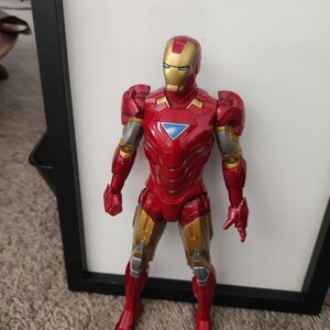 May include: An Iron Man action figure featuring a metallic red and gold suit with a triangular blue chest piece. The character is depicted in a standing position with articulated limbs, a detailed helmet, and silver accents on the arms and legs.