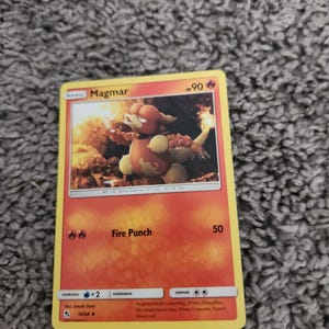 May include: A Pokémon trading card featuring Magmar, a fiery creature with orange and yellow coloring. The card has the text "Magmar" and "Fire Punch" with a power level of 50. The card is set against a fiery background.