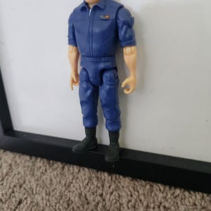 K&ouml;nnte beinhalten: Eine blaue und orangefarbene Actionfigur eines Mannes, der einen blauen Overall mit Rei&szlig;verschluss und einem Aufn&auml;her auf der Brust tr&auml;gt. Die Figur hat braunes Haar und steht auf einer schwarzen Oberfl&auml;che.