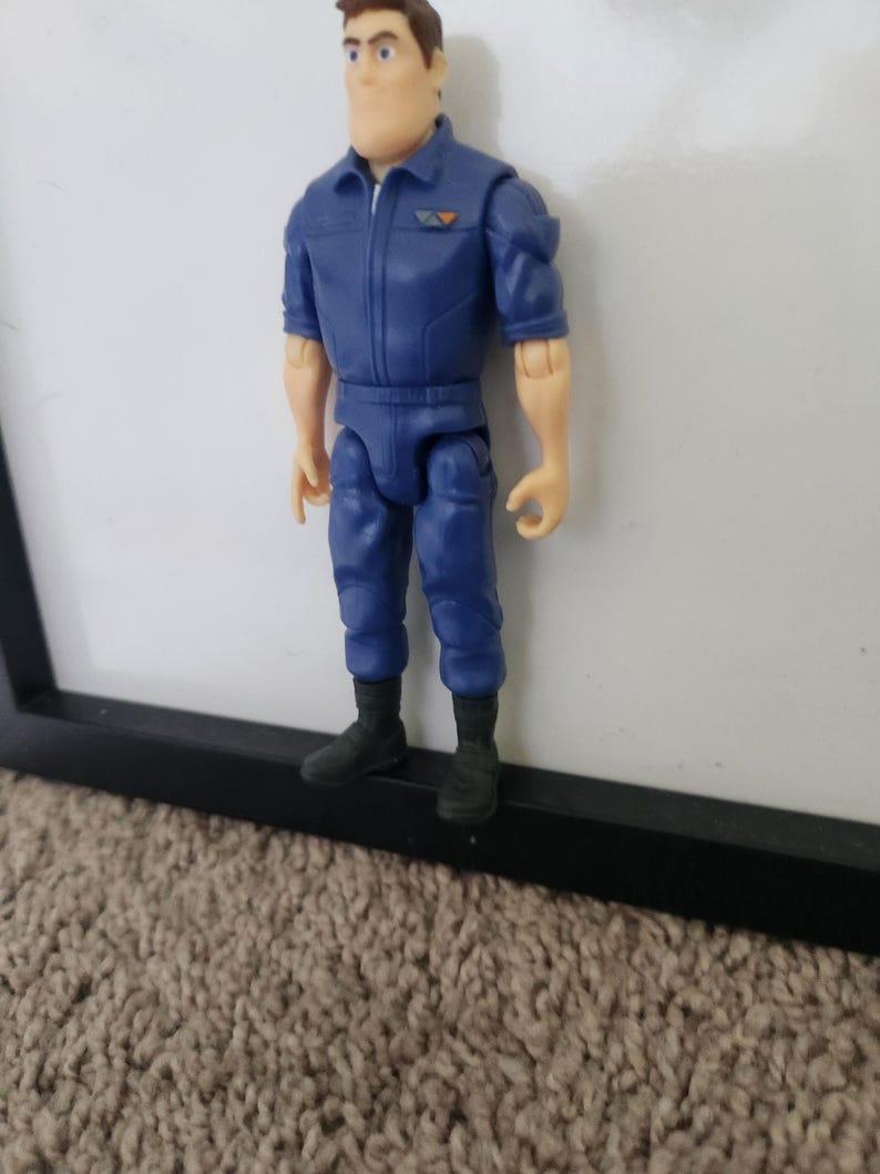 K&ouml;nnte beinhalten: Eine blaue und orangefarbene Actionfigur eines Mannes, der einen blauen Overall mit Rei&szlig;verschluss und einem Aufn&auml;her auf der Brust tr&auml;gt. Die Figur hat braunes Haar und steht auf einer schwarzen Oberfl&auml;che.