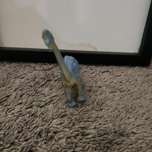 May include: A small, blue-grey plastic dinosaur figurine with a long neck and tail, standing on four legs. The figure has a smooth texture and is depicted in a standing position, showing the front view of the head and the length of the neck.